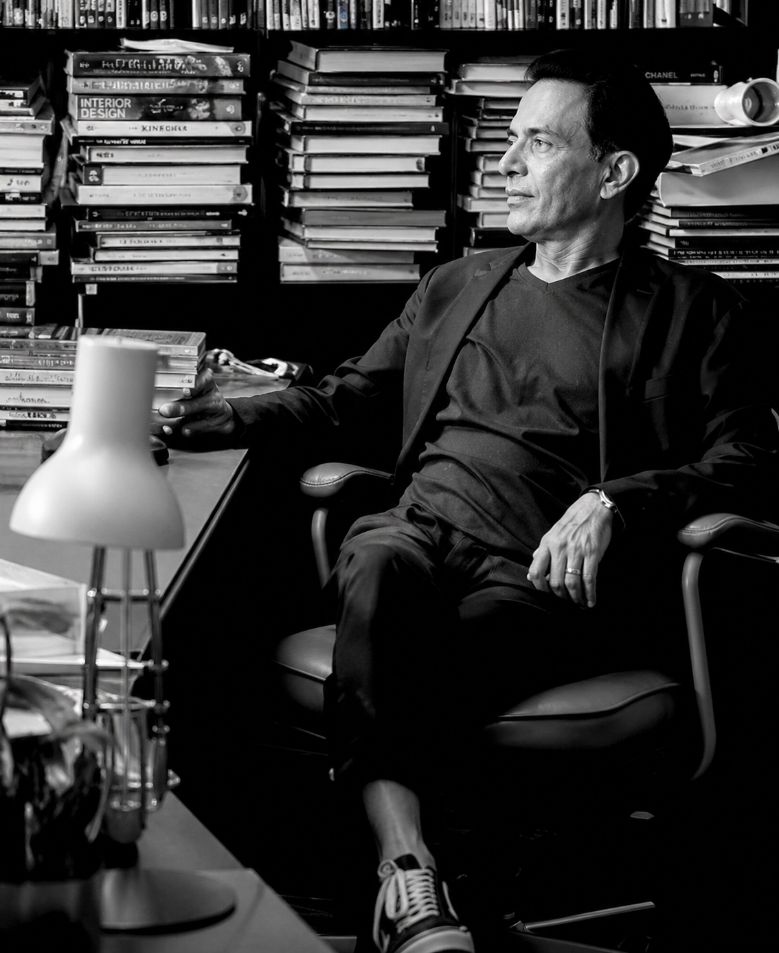 Man in a dark blazer sits casually in an office chair at a cluttered desk, with tall shelves of books behind him (black-and-white).