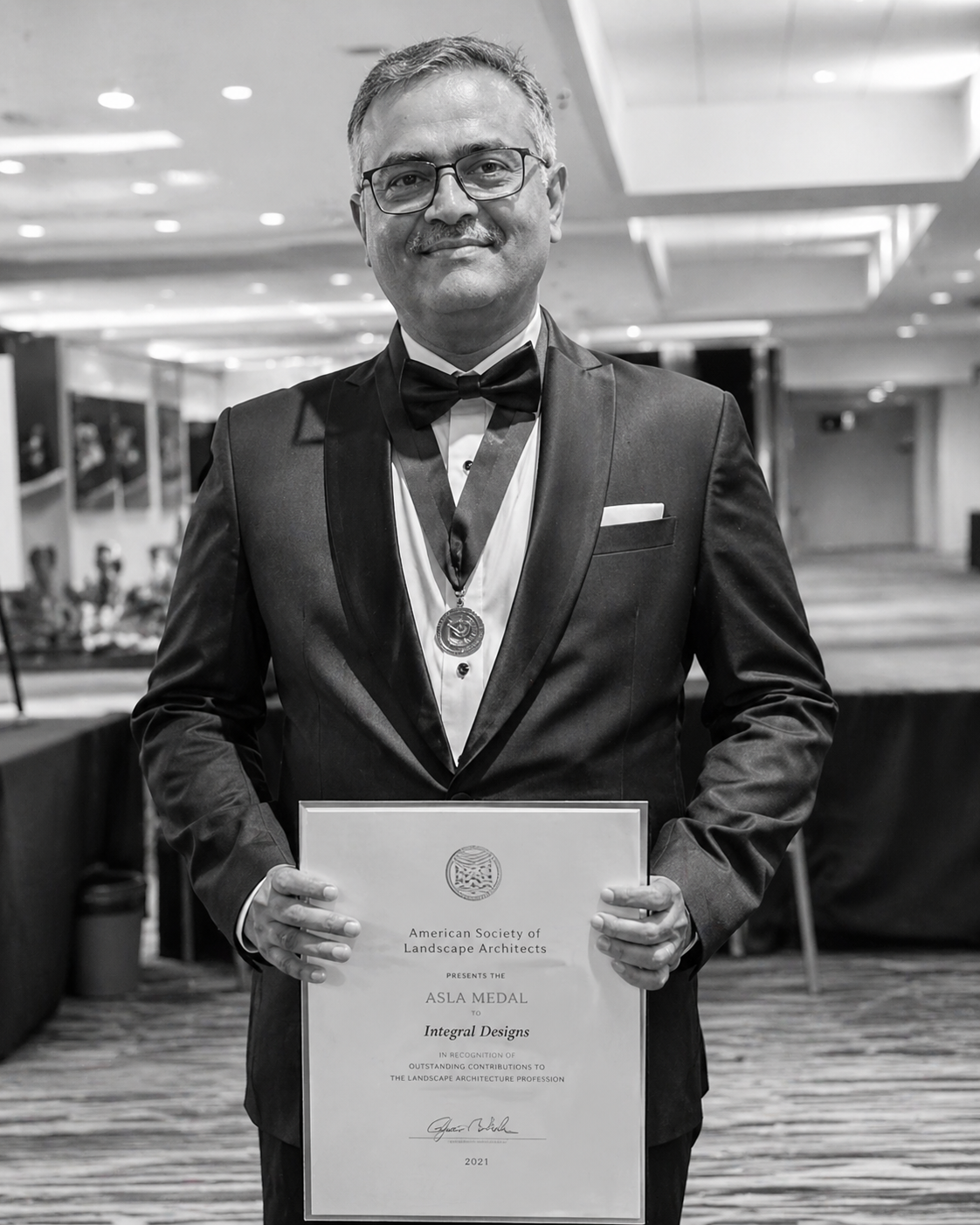 Smiling man in a tuxedo and bow tie holding an award certificate with a medal hanging around his neck in a conference setting.