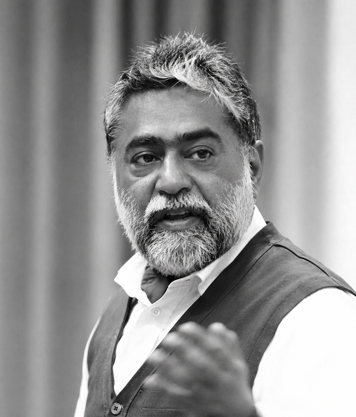 Portrait of a bearded man in a white shirt and dark vest, mid-speech, gesturing with his hand.